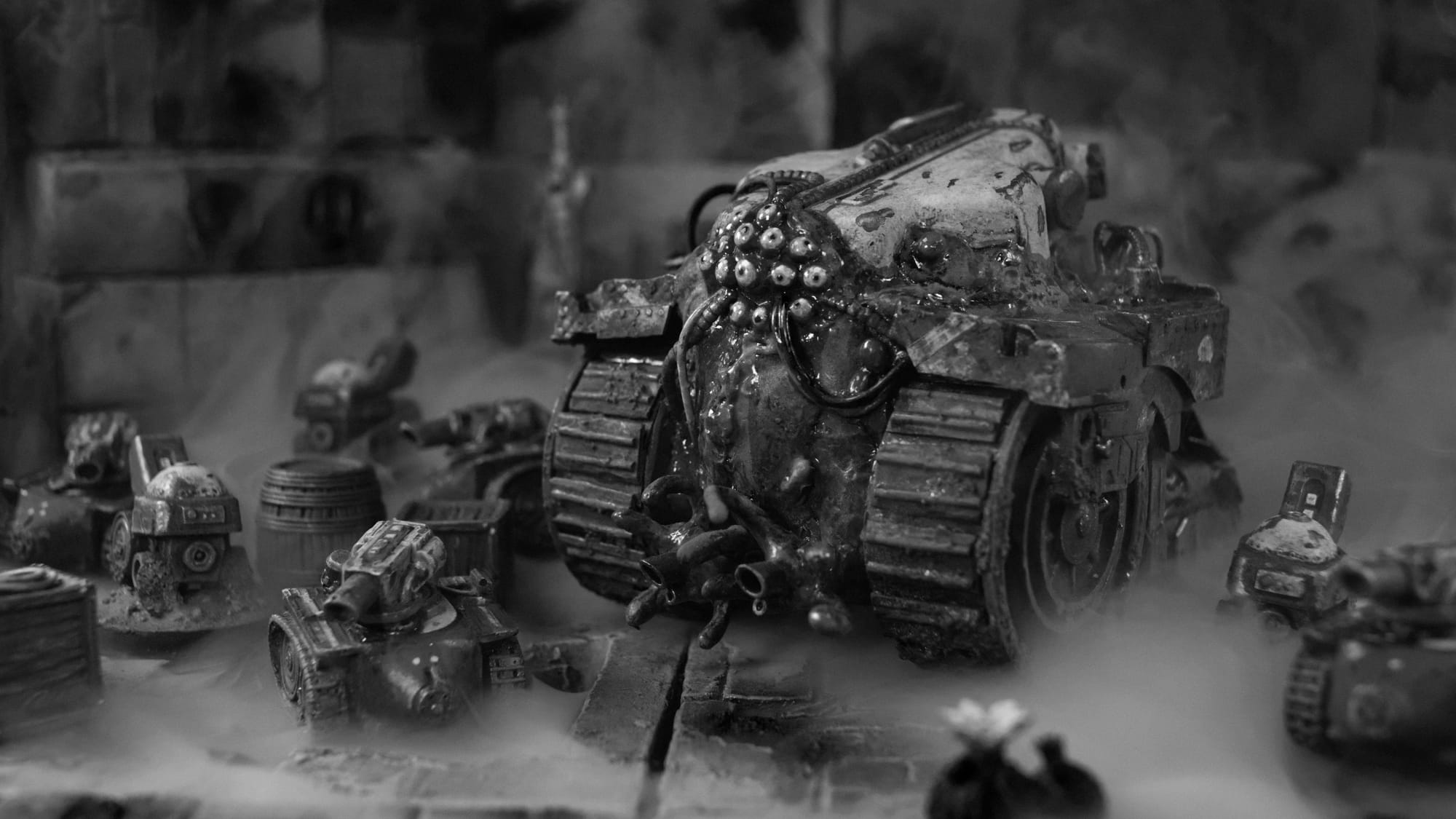 Black and white photo of a big gruesome biomechanical tank with lots of eyes surrounded by numerous smaller vehicles in a misty cityscape.